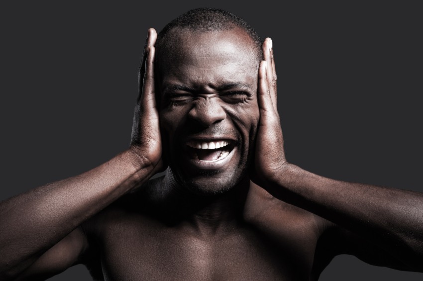 Too loud sound. Portrait of young shirtless African man covering ears with hands and shouting while standing against grey background